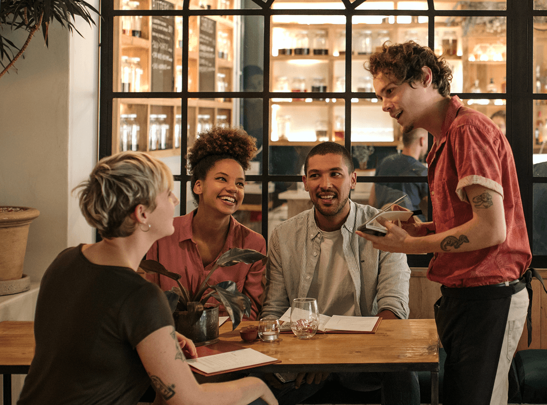 Um grupo de amigos pratica um novo idioma que aprenderam enquanto fazem um pedido a um garçom em um café elegante.