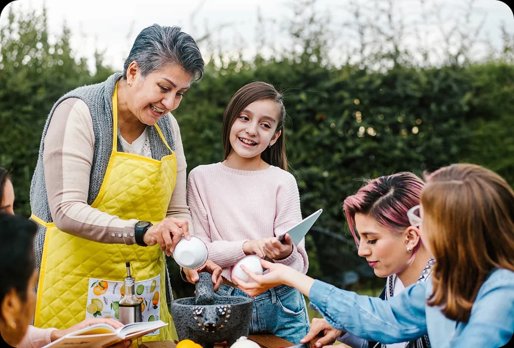 Um adulto ensina crianças a preparar ingredientes com um almofariz e um pilão durante uma atividade prática de aprendizagem cultural ao ar livre.