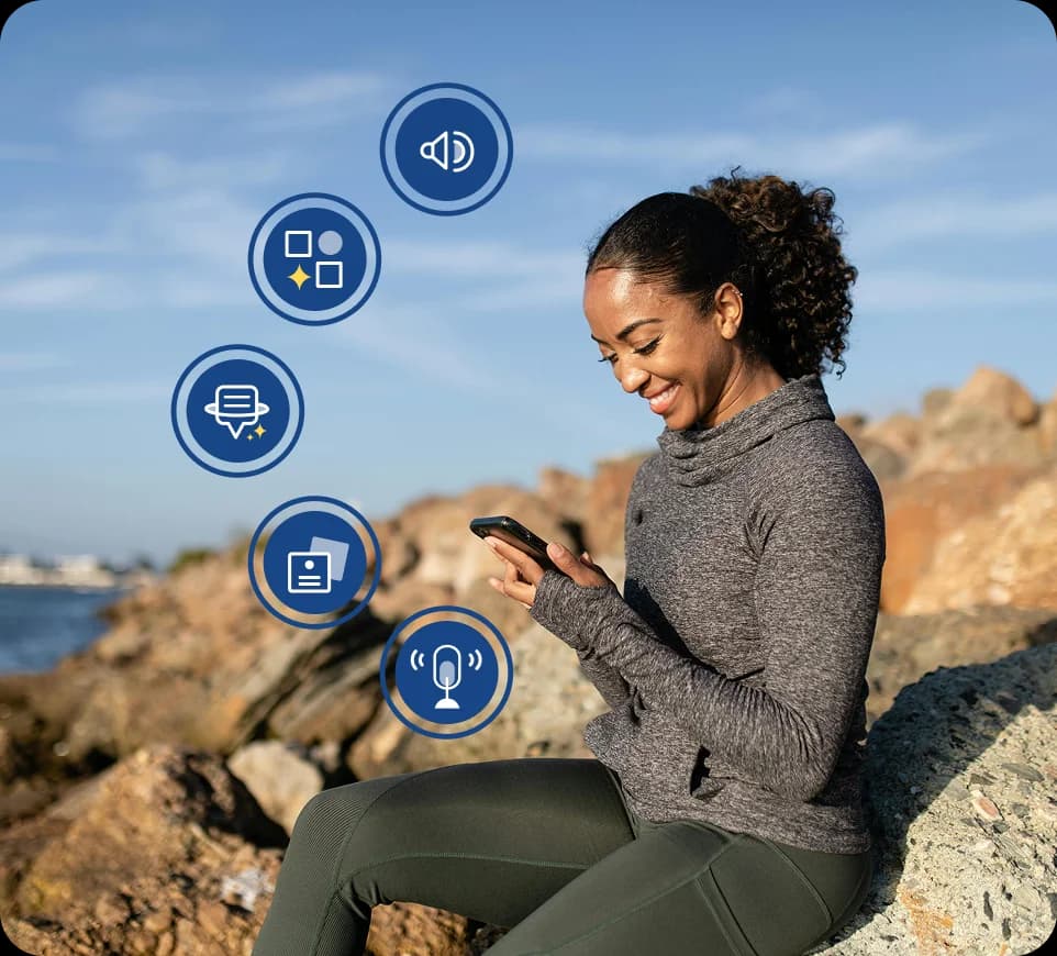 Uma mulher sentada ao ar livre em rochas perto da água, sorrindo e olhando para o seu celular, com ícones de funcionalidades do Rosetta Stone Sapphire sobrepostos à sua volta.